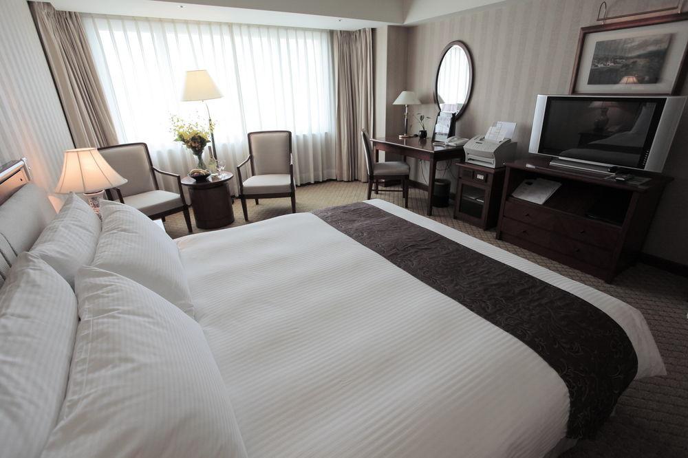 Evergreen Laurel Hotel - Tchaj-čung Exteriér fotografie The photo shows a well-furnished hotel room. In the foreground, there is a large bed with white bedding and a dark-colored throw at the foot. Beside the bed, there is a bedside lamp. 

In the background, natural light filters through large windows co
