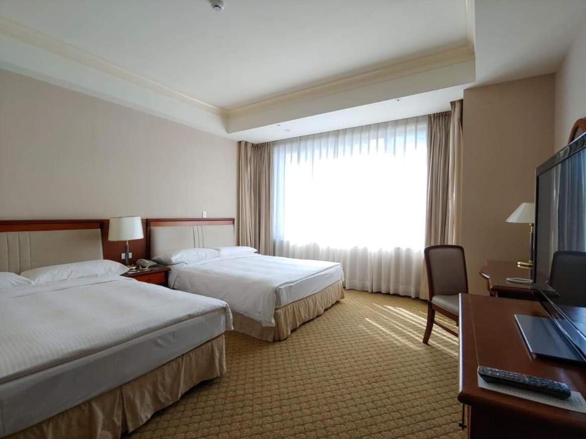 Evergreen Laurel Hotel - Tchaj-čung Exteriér fotografie The image shows a hotel room featuring two single beds with white bedding. There is a large window with sheer curtains letting in natural light, and the walls are painted in neutral tones. A wooden desk with a chair is positioned near the window, and