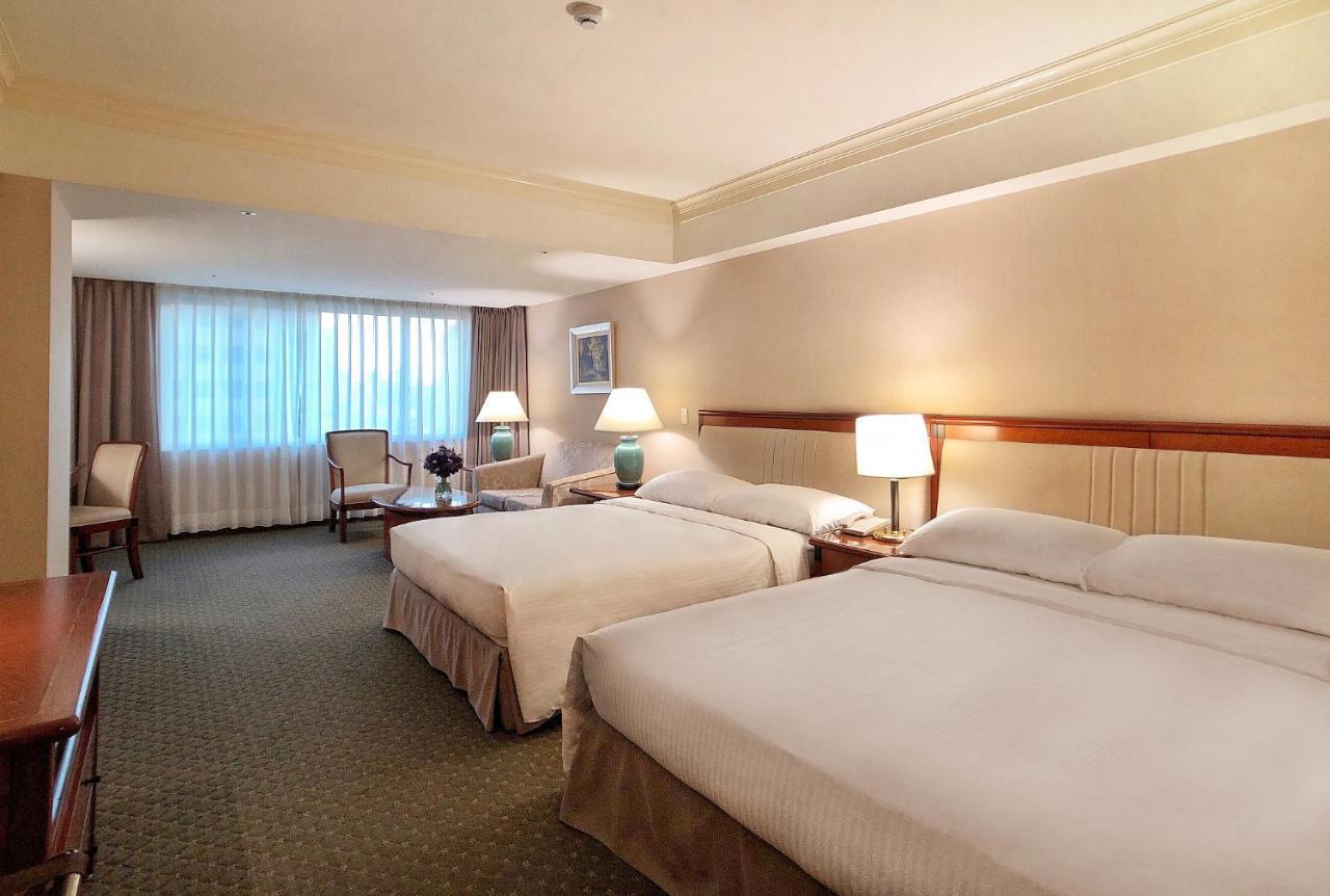 Evergreen Laurel Hotel - Tchaj-čung Exteriér fotografie The photo shows a hotel room featuring two queen-sized beds with white linens. The room is well-lit and has a warm, neutral color palette. There are bedside lamps on each side of the beds and a small table. To the side, there's a comfortable armchair