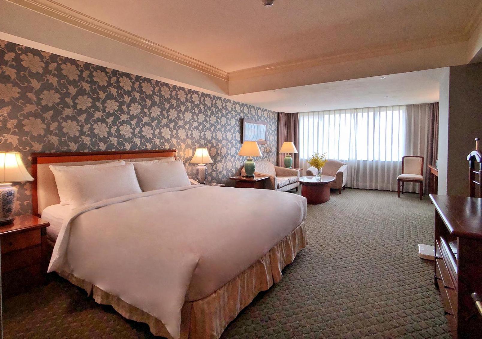 Evergreen Laurel Hotel - Tchaj-čung Exteriér fotografie The photo shows a hotel room with a spacious and comfortable layout. It features a large bed with white bedding, placed against a decorative wallpapered wall. On either side of the bed, there are lamps providing soft lighting. In addition to the bed,