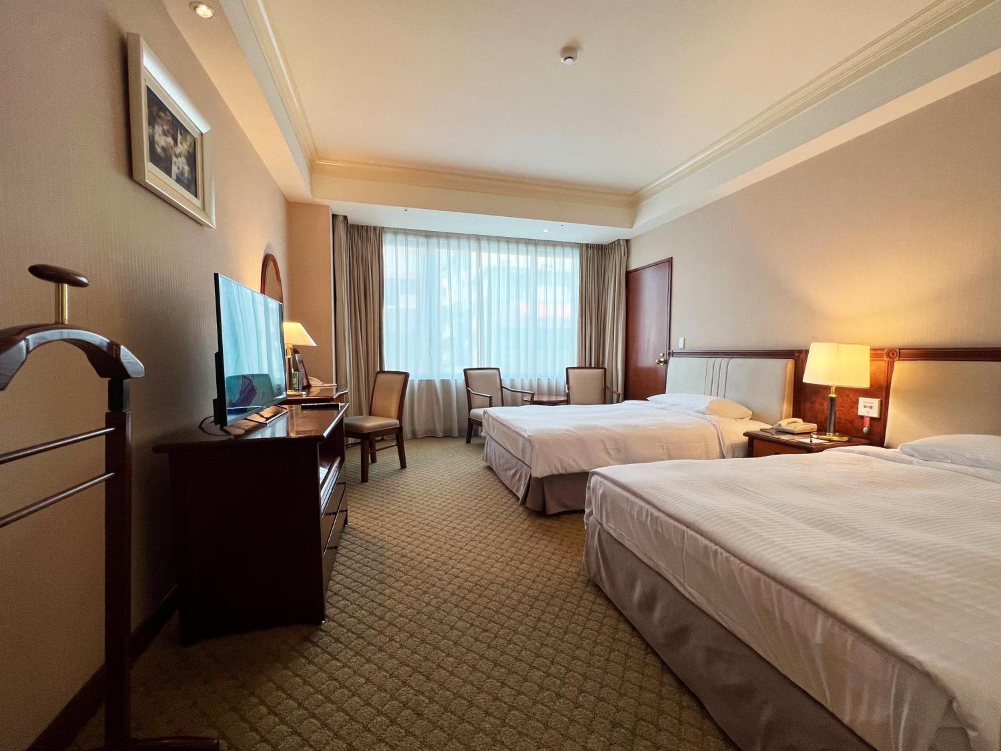 Evergreen Laurel Hotel - Tchaj-čung Exteriér fotografie The photo shows a hotel room featuring two twin beds placed parallel to each other. The room has a warm color palette and includes a large window with sheer curtains allowing natural light to illuminate the space. There is a wooden desk and two chair