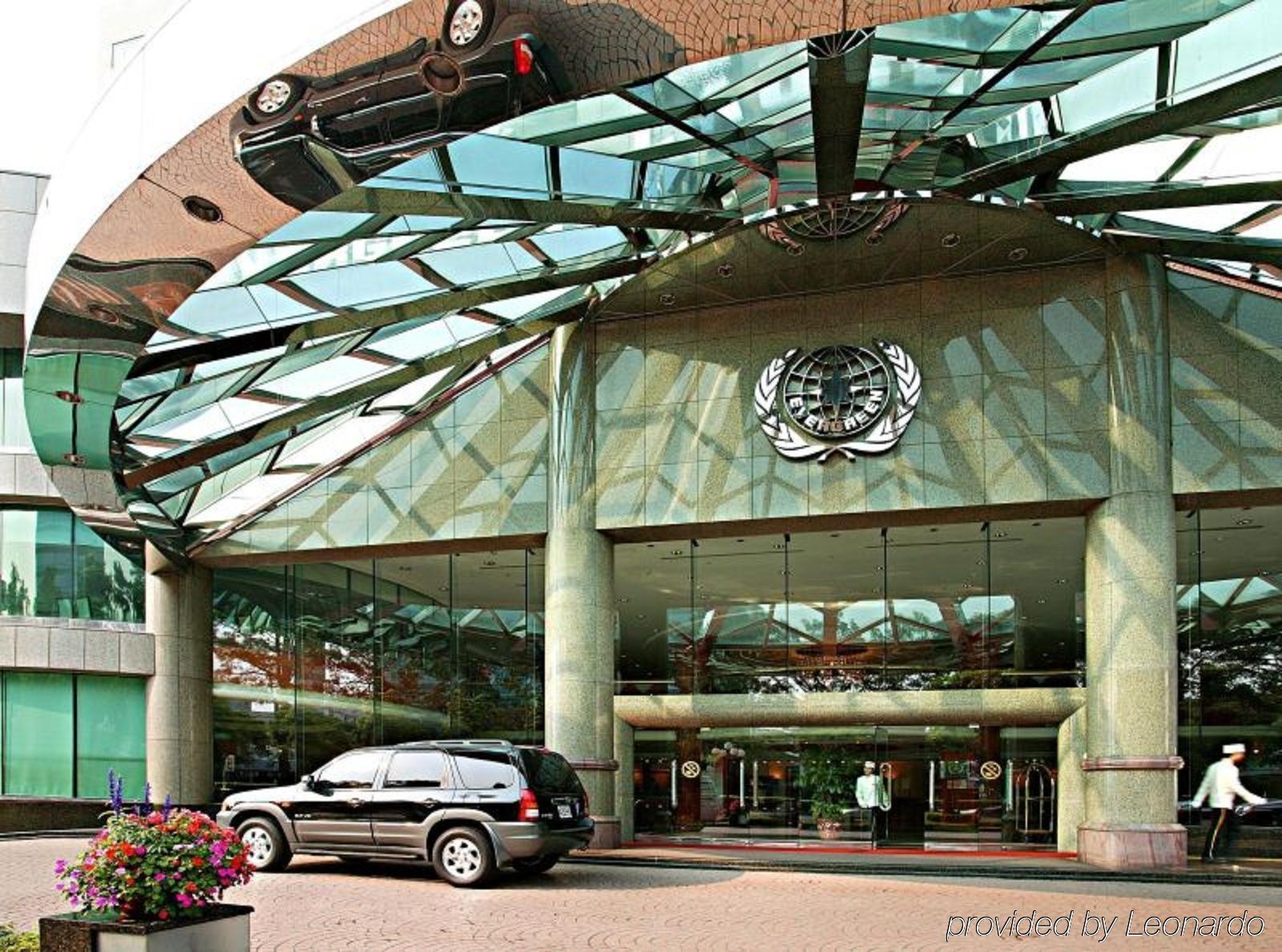 Evergreen Laurel Hotel - Tchaj-čung Exteriér fotografie The photo shows the entrance of a modern building characterized by its glass and metal architecture. The structure features a large, domed roof that reflects light, with a distinctive emblem or logo displayed prominently at the center. In front of th