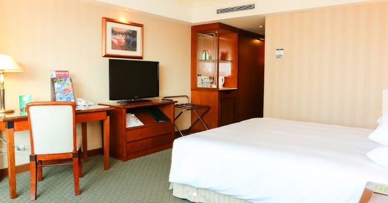 Evergreen Laurel Hotel - Tchaj-čung Exteriér fotografie The photo shows a hotel room featuring a neatly made bed with a white blanket. On one side, there is a wooden desk with a chair, and a flat-screen television is mounted nearby. An accent cabinet is visible, displaying various items. The walls are pai