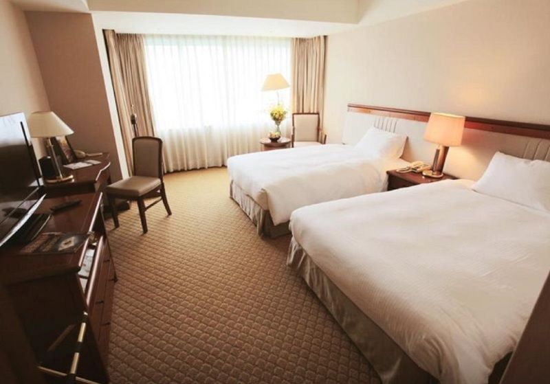 Evergreen Laurel Hotel - Tchaj-čung Exteriér fotografie The photo shows a hotel room featuring two twin beds with white bedding. There's a chair positioned near one of the beds and a desk with a computer setup against one wall. Large windows allow natural light to enter the room, and light-colored curtain