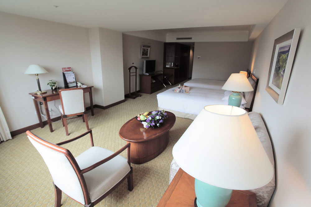 Evergreen Laurel Hotel - Tchaj-čung Exteriér fotografie The photo shows an interior view of a hotel room. It features two beds, a coffee table with a small decorative arrangement of flowers, and a comfortable chair. There's a desk and a lamp, along with another larger lamp on the table. In the background,