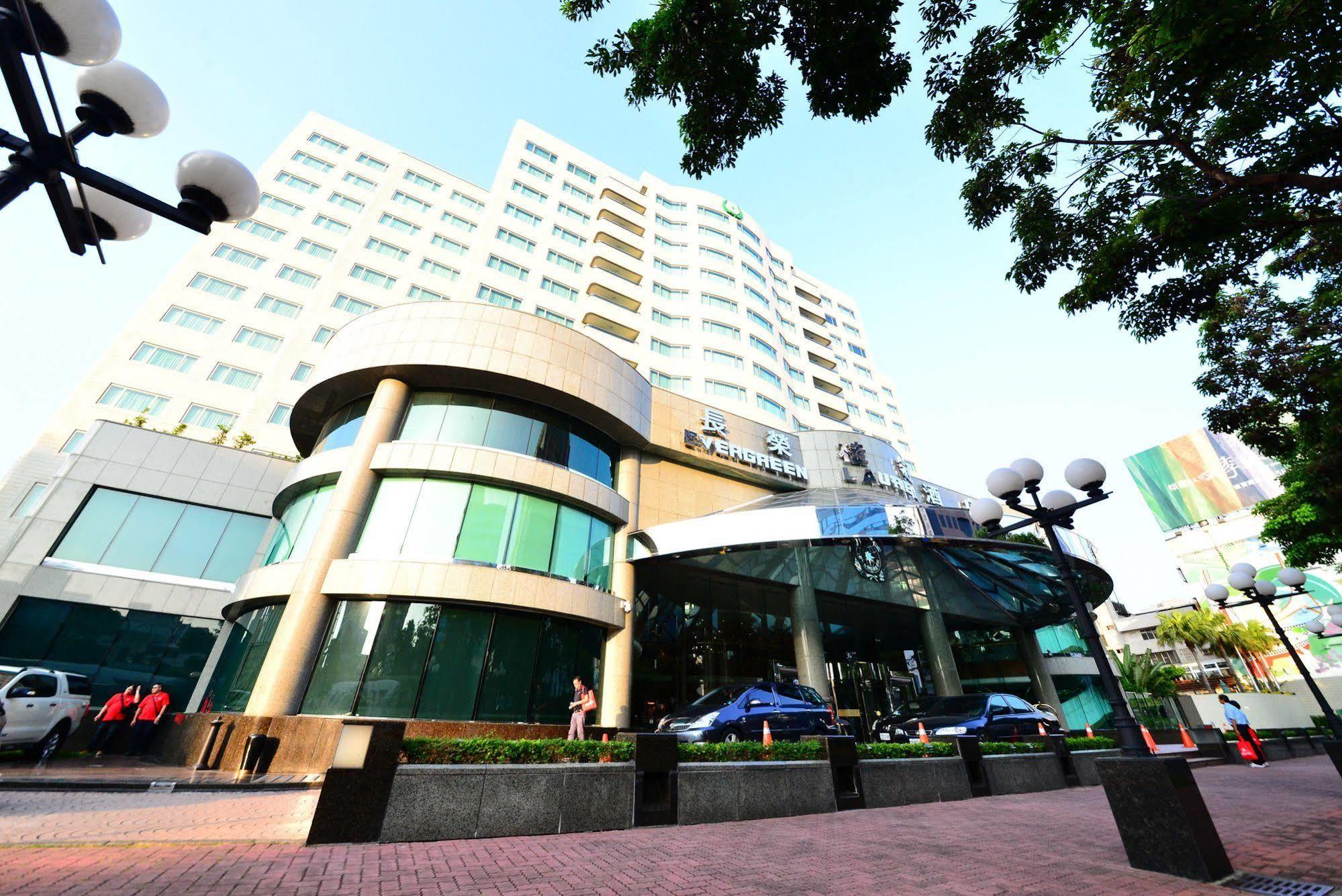 Evergreen Laurel Hotel - Tchaj-čung Exteriér fotografie The photo shows a modern multi-story building, which appears to be a hotel or commercial establishment. The structure has a rounded glass entrance with a distinctive architectural design, featuring large windows. The building is surrounded by greener