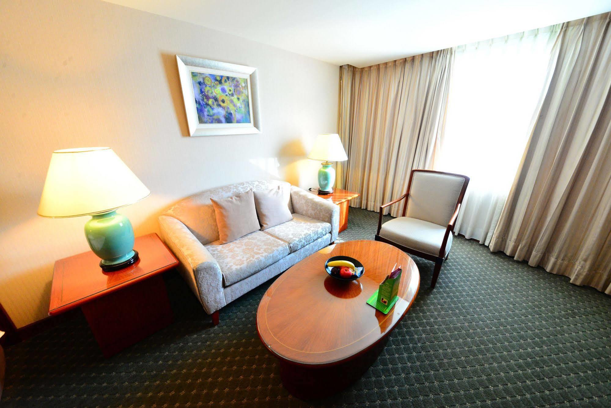 Evergreen Laurel Hotel - Tchaj-čung Exteriér fotografie The photo shows a cozy living space, likely a hotel room or a lounge area. It features a light-colored sofa with cushions, a wooden coffee table in the center, and a comfortable armchair beside it. There are two lamps with turquoise bases on side tab