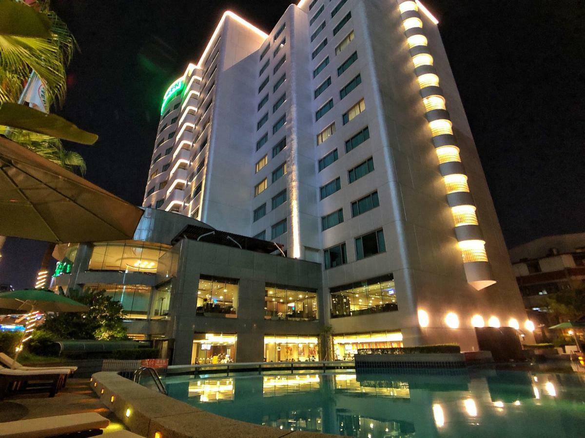 Evergreen Laurel Hotel - Tchaj-čung Exteriér fotografie The photo shows a tall hotel building at night, illuminated by lights. The architecture appears modern, with several floors and large windows. In the foreground, there is a swimming pool that reflects the lights from the building, creating a serene a