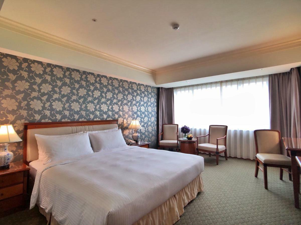 Evergreen Laurel Hotel - Tchaj-čung Exteriér fotografie The photo shows a hotel room that features a large bed with a white bedspread positioned against a decorative floral wallpaper. There are two bedside lamps on wooden nightstands flanking the bed. To the side of the room, there are two chairs and a sm