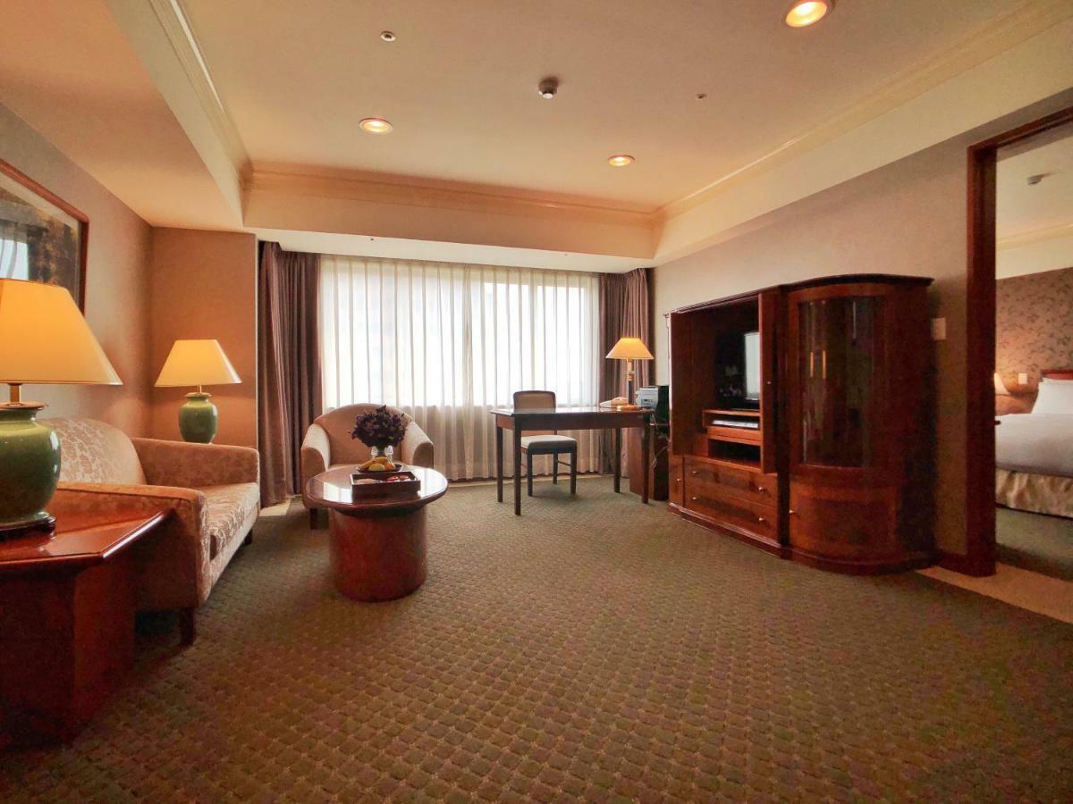 Evergreen Laurel Hotel - Tchaj-čung Exteriér fotografie The photo shows a cozy hotel suite or living area. There is a sofa and an armchair arranged around a small coffee table. A desk with a chair is positioned near a large window covered with sheer curtains that allow natural light to enter the room. Two