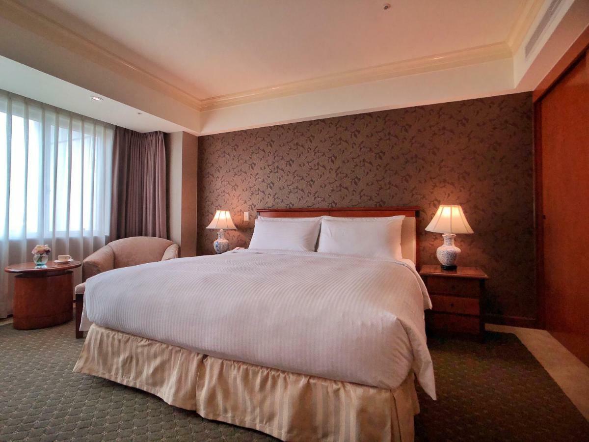 Evergreen Laurel Hotel - Tchaj-čung Exteriér fotografie The photo shows a neatly arranged hotel room featuring a large bed with white linens and a light-colored bed skirt. On either side of the bed, there are small wooden nightstands with lamps. To the left of the bed, there's a comfortable chair and a ro
