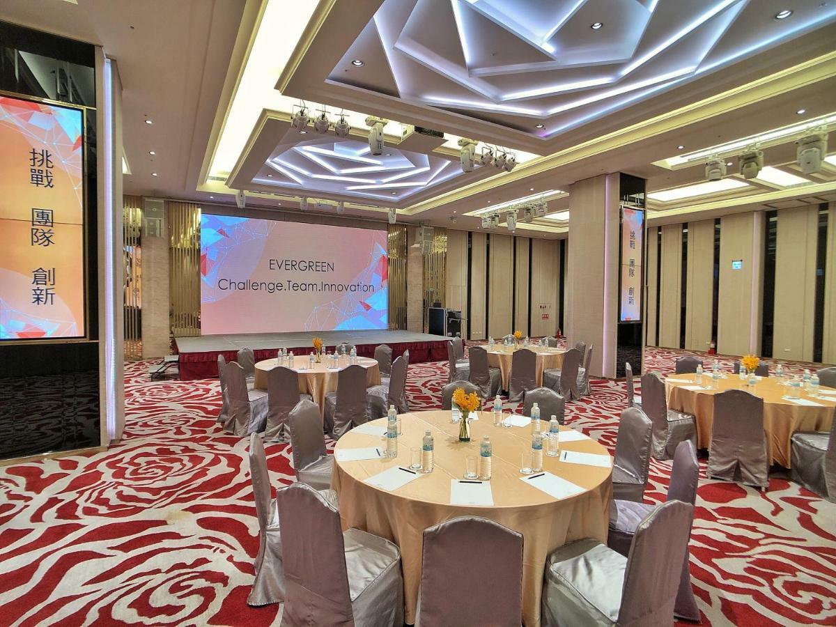 Evergreen Laurel Hotel - Tchaj-čung Exteriér fotografie The photo shows a conference or event space set up for a gathering. The room features several round tables with light-colored tablecloths and chairs, each with decoration, possibly for dining or discussion. A large screen displays the text "EVERGREEN