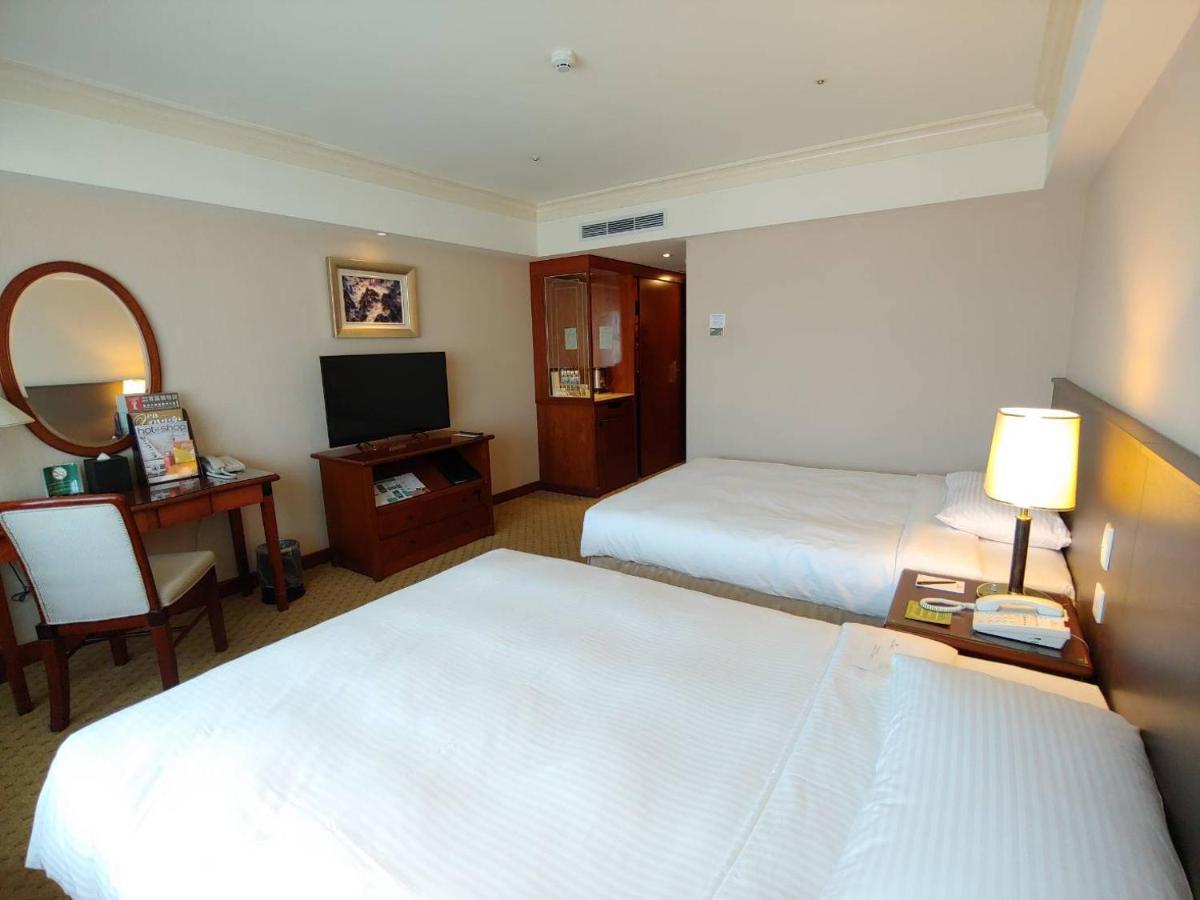 Evergreen Laurel Hotel - Tchaj-čung Exteriér fotografie The photo shows a hotel room with two beds, neatly made with white linens. There is a desk and chair in one corner, accompanied by a flat-screen TV mounted on the wall. A large mirror and a wooden cabinet are also visible. The room is illuminated by 