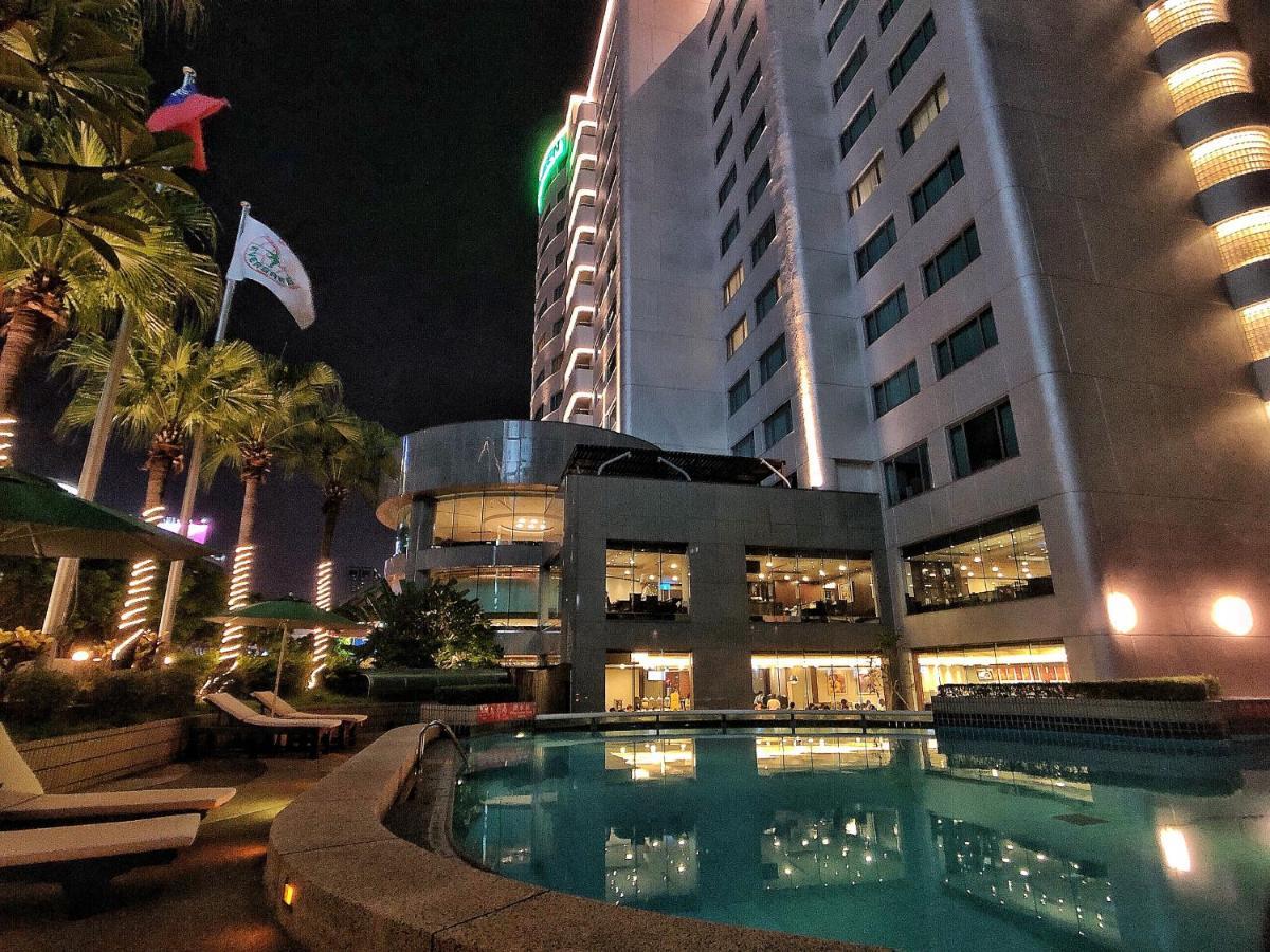 Evergreen Laurel Hotel - Tchaj-čung Exteriér fotografie The photo depicts a modern hotel or building at night, featuring a sleek facade illuminated by lights. In the foreground, there is a swimming pool that reflects the surrounding lights and palm trees. There are lounge chairs near the pool, and the atm